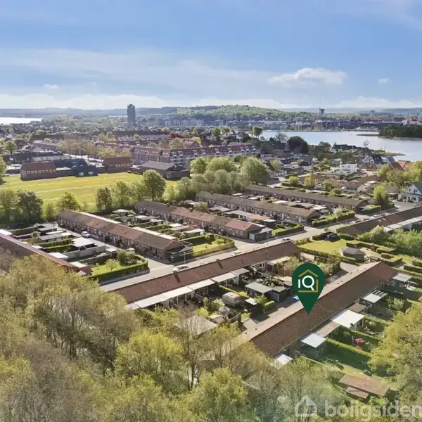 Rækkehuse ses fra luften omgivet af træer og grønne områder, med en sø i baggrunden. En markør identificerer en specifik bygning. Byen og landskabet strækker sig mod horisonten.