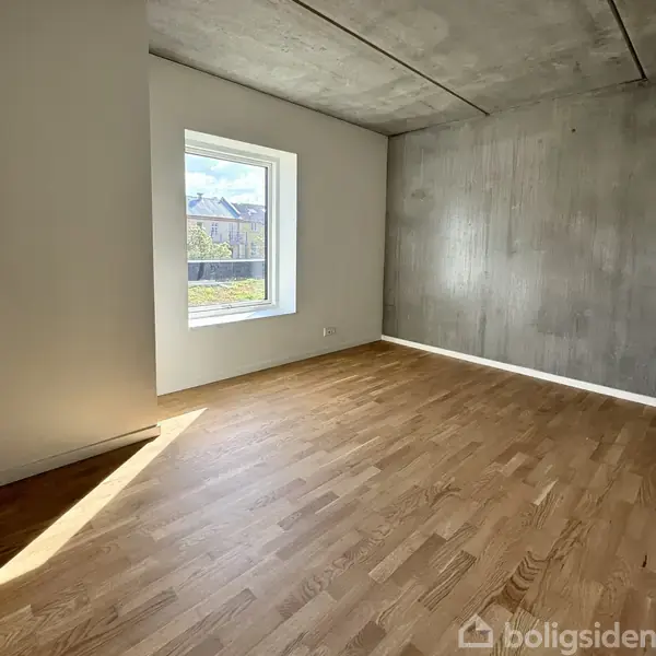 Et tomt rum med trægulv og hvidmalede vægge. Et vindue til venstre slipper dagslys ind, og en struktureret væg til højre giver rummet karakter. Udsigt til bygninger udenfor.