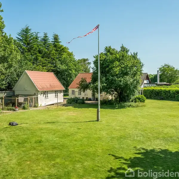Et hus med rødt tag står ved siden af et mindre udhus på en velholdt græsplæne. En flagstang med flag er centralt placeret i haven, omgivet af træer og hække.
