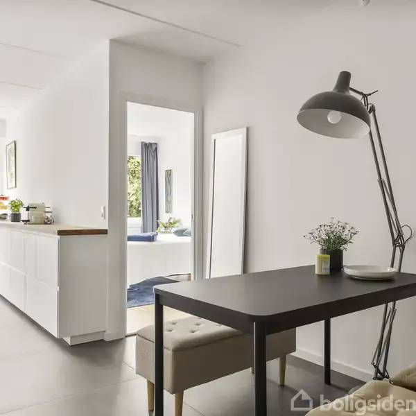 Et spisebord med to beige bænke og en stor, sort gulvlampe i en moderne indrettet gang. I baggrunden ses et lyst køkken med vinduer og grøn udsigt.