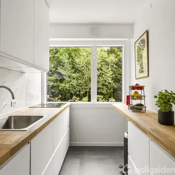 Et lyst køkken med hvide skabe og en træbordplade. En vask og kogeplade er integreret. Store vinduer åbner op mod en grøn udsigt. En plante og krydderirack pryder bordet.