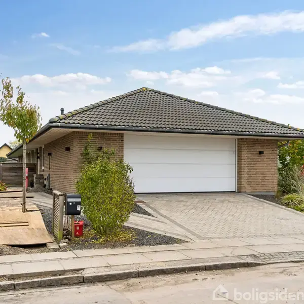 En étplans villa i mursten med grå tagsten og en stor hvid garageport. Omgivet af en indkørsel i fliser, grønne buske og træer på en stille villavej.