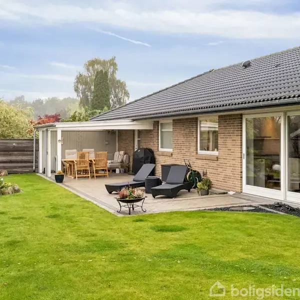 En murstensvilla med gråt tegltag har en bred hvid garageport. Foran er der en indkørsel med belægningssten, omkranset af buske og træer i et velplejet haveområde.