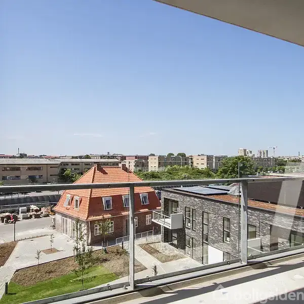 En altan med glasrækværk giver udsigt over en moderne bydel med forskellige bygninger, herunder røde tegltag og grå facade. Solrig atmosfære, blå himmel.