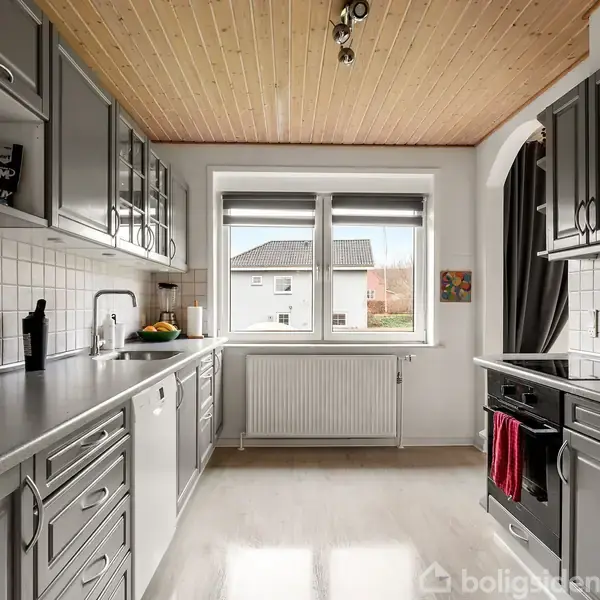 Køkken med grå skabe og skuffer langs væggene, en hvide kakkelvæg, og en stor vindue med udsigt til et hus udenfor. Loft i træ.