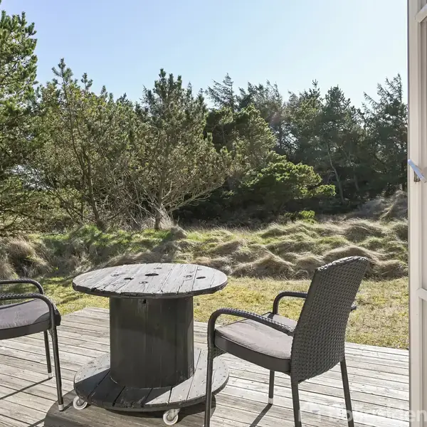To stole omkring et hjemmelavet bord af en kabeltromle på en træterrasse. I baggrunden ses en skovklædt klitlandskab under en klar himmel.