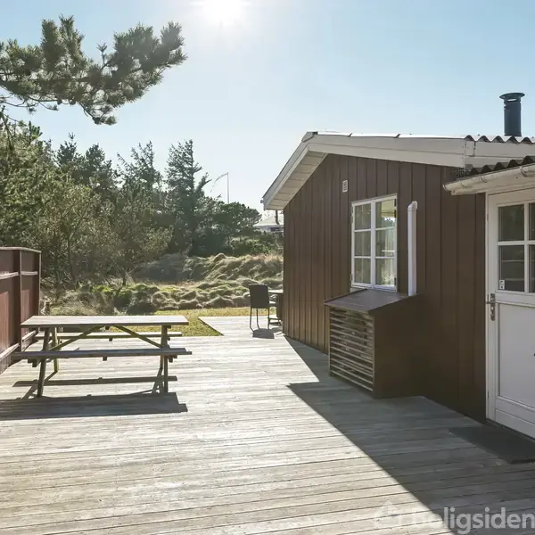 Træterrasse ved et rødt sommerhus med en hvid dør. Et bord-bænkesæt står til venstre, omkranset af en lav, rød træhegn. Omgivet af træer og buske.