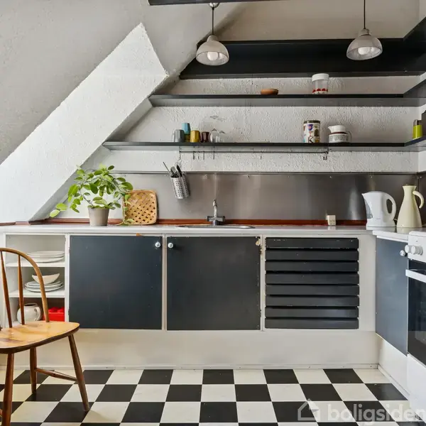 Køkken med sorte og hvide fliser. Hylder med skåle og dåser. Køkkenbord med planter, brødkasse, elkedel og stikkontakt. Træstol ved vindue. Loftlamper over bordet.