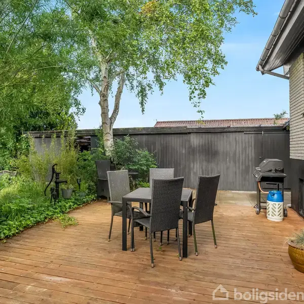 En terrasse med et bord og seks stole står på et trædæk omringet af grønne planter. En grill er placeret tæt ved huset. En birketræ ses i baggrunden.