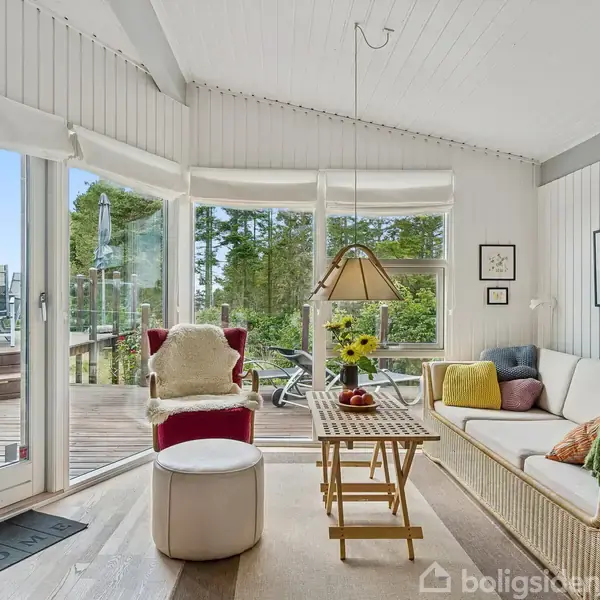 En lys stue med en sofa, hvor der ligger farverige puder. Et bord med en vase med blomster står foran. Store vinduer viser en terrasse med møbler udenfor.