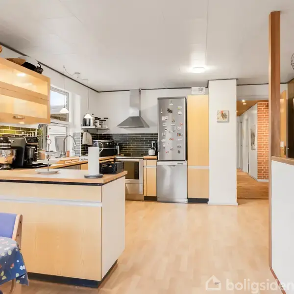 Et moderne køkken med lyse trægulve og træskabe. Induktionskomfur på bordet, køleskab i rustfrit stål, sorte flisevægge. Til højre ses en gang med røde murstensvægge.