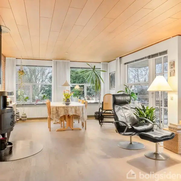 En lounge stol står i en lys, luftigt indrettet stue med store vinduer, der lukker naturligt lys ind. En kamin og spisestue ses i baggrunden, omgivet af grønne planter.