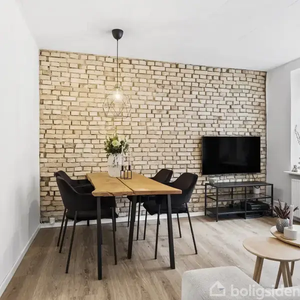 Stue med grå sofa og runde sofaborde foran en væg med kunsttryk. Ved siden af står et spisebord med sorte stole og blomster. Murstensvæg i baggrunden.