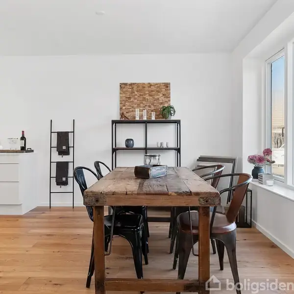 Et rustikt spisebord med sorte stole står i et lyst køkken-alrum. Bordet er dækket med en avis, og ved siden af er en hvid skænk og hylder med dekorationer. En stor vindueskarm med blomster skaber lys.