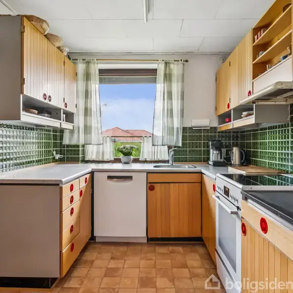 Et køkken med træpaneler på skabene udfører opgaven i et retro-design. Grønne flisevægge omgiver rummet, mens vinduet viser et udendørs landskab med blå himmel og tage.