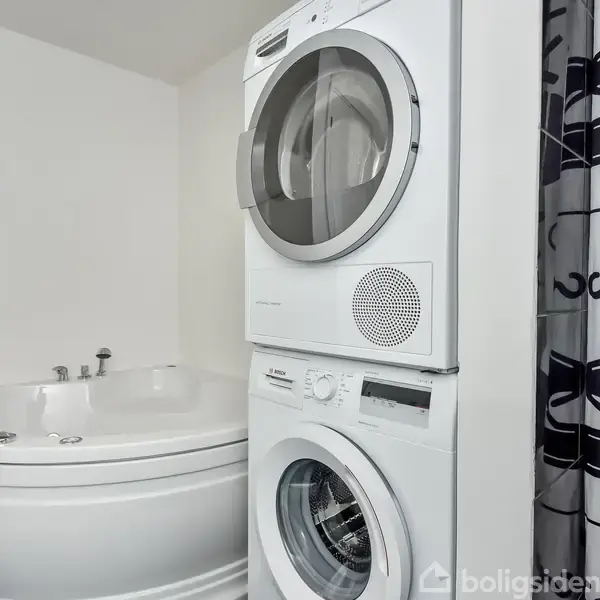 En stabel af en vaskemaskine og en tørretumbler står i et badeværelse ved siden af et hjørnebadekar og et mønstret badeforhæng.