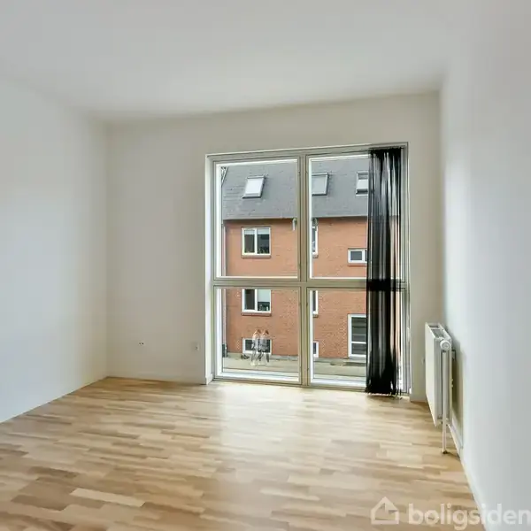 Et tomt rum med trægulv, sollys fra et stort vindue. Udenfor ses røde murstensbygninger. Rummet er hvidmalet med en radiator ved væggen.