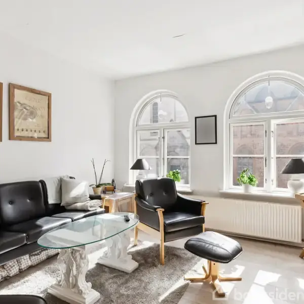 Sort lædermøblement står i en stue med et rundt glasbord, flankeret af to billeder på væggen. Store buede vinduer lader sollys ind, og planter er placeret på vindueskarmen.