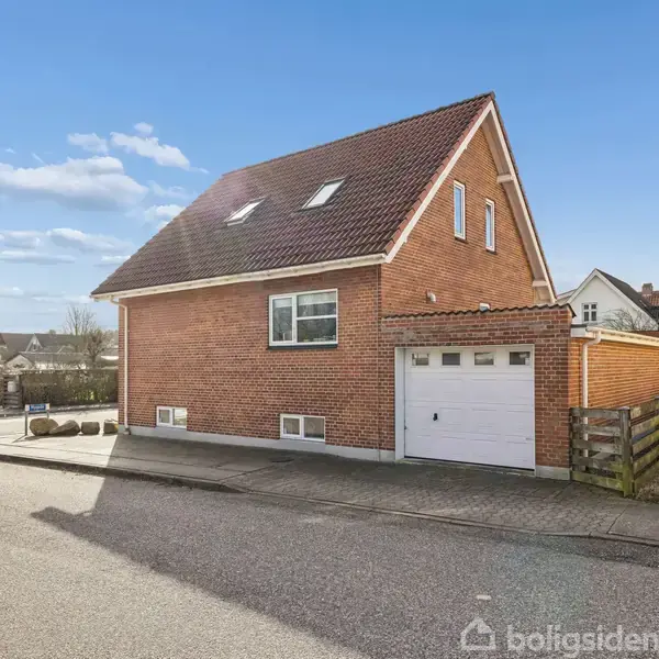Rødt murstenshus med garage, stående stille på en villavej i et beboelseskvarter, omgivet af andre huse og biler under en blå himmel med få skyer.