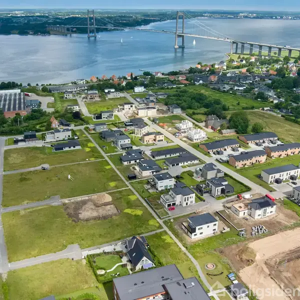 Nybyggeri af huse ligger langs grønne områder, tæt på en bro der krydser en bred vandstrøm. Området er omgivet af natur og småbyer.