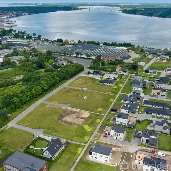 Boligområde med nye huse ligger ved en flod med en bro i baggrunden. Området er omgivet af grønne områder og en by i nærheden. Tekst ignoreret: "Boligsiden".