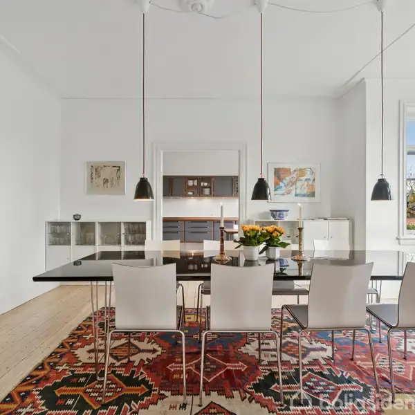 Et spisebord med otte stole omringet af et farverigt tæppe. Bordet er dekoreret med en vase blomster. Rummet er lyst med hvide vægge og store vinduer.