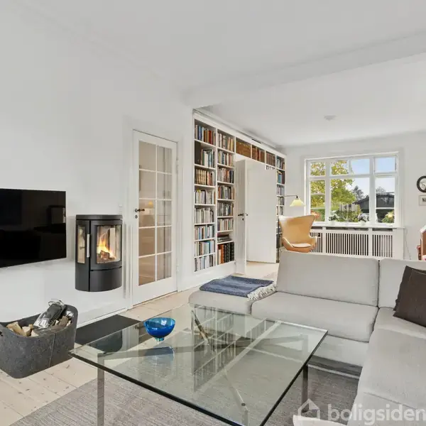 En stue med en grå sofa placeret omkring et glasbord. Pejsen er tændt, og en bogreol fylder bagvæggen. Store vinduer giver udsigt til en have. Fjernsynet hænger på væggen.