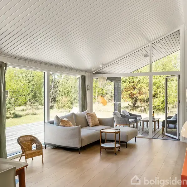 Stue med moderne pejs, to sofaer og sofabord, orienteret mod store glasdøre, der åbner til en terrasse. Skoven ses udenfor gennem de store vinduer.
