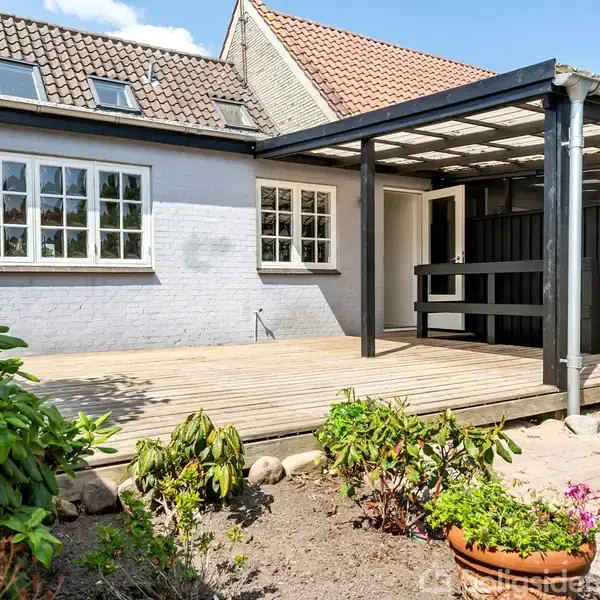 En terrasse udendørs med en træveranda dækket af en transparent tagkonstruktion. Terrassen er omgivet af et hus med lyse mursten, og der er potteplanter og buske omkring.
