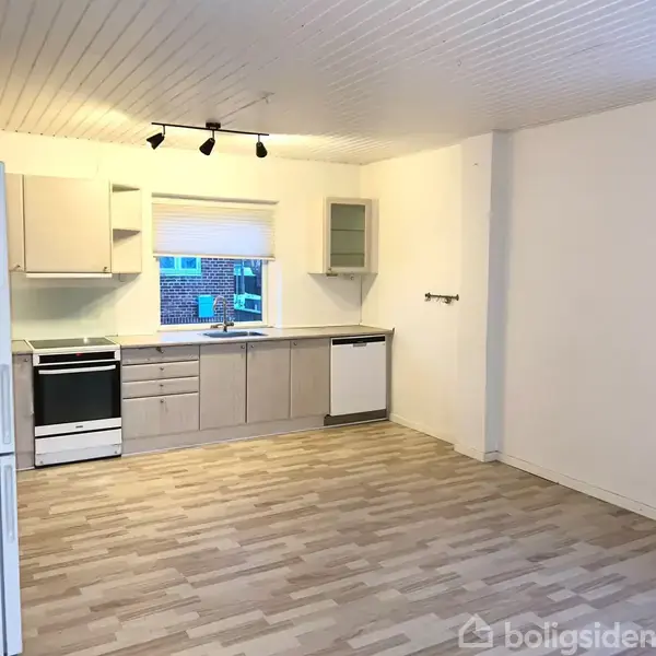 Et køkken indeholder hvidt køleskab, komfur og opvaskemaskine. Trægulvet er lyst, og rummet har hvide vægge og loft med indbygget belysning. Et vindue viser murstensbygning udenfor.