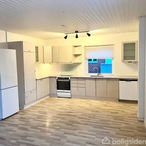 Køkken med hvide skabe og apparater som køleskab og komfur, placeret i et rummeligt rum med trægulv. En stor vindue ved vasken giver udsigt til murstensbygning udenfor.