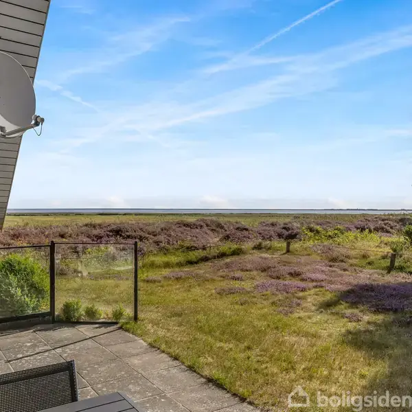 Terrasse med stole og bord står stille; den vender mod et åbent landskab med lilla lyng og græs. En stor parabol hænger fra taget ved siden af en rolig, blå himmel.