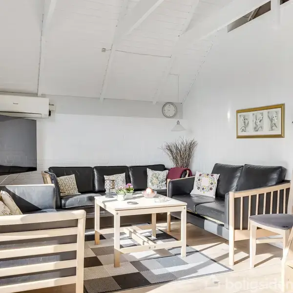 Stue med sort sofa og sofaborde i træ. Solskin gennem store vinduer oplyser rummet med lyse træmøbler og dekorative puder. En stige fører opad, mens vægkunst hænger til højre.