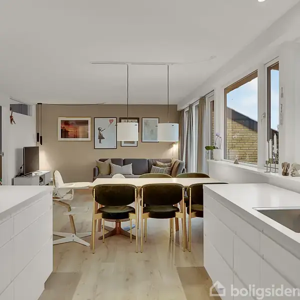 Køkken og stue i åben planløsning; hvidt interiør med spisebord og stole; stor vinduesrække på højre side med udsigt til have; sofa og billeder på væggen i baggrunden.