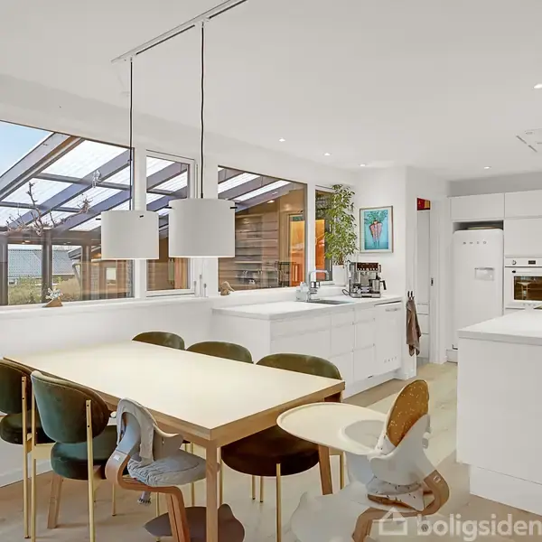 Et moderne køkken med et hvidt spisebord omgivet af stole, beliggende i et lyst rum. Store vinduer giver udsigt til en overdækket terrasse. Peberkværne og kaffemaskine på bordet.
