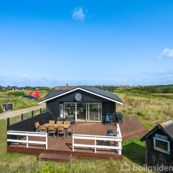 Sort sommerhus med stor træterrasse, hvor der står et bord med stole. Huset ligger i en grønt og åbent landskab med enkelte andre huse i nærheden under en blå himmel.