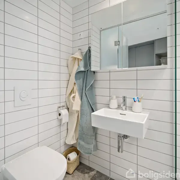 Toilet først med håndvask til højre, dekoreret med tandbørsteholder og sæbedispenser. Flisebelagte vægge og gulv, med håndklæder hængende og papirkurv under vask i et moderne badeværelse.