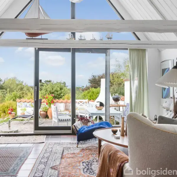 Stue med skydedøre åbner ud til en terrasse med udsigt over en have. Rummet har lyse trælofter, møbleret med en sofa, stole og bogreol. Sollys oplyser interiøret.