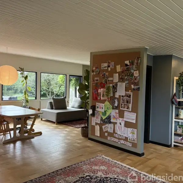 Spisebord med stole står ved store vinduer, der giver udsigt til haven. Ved siden af er en sofa. På en søjle hænger en opslagstavle fuld af noter og billeder.