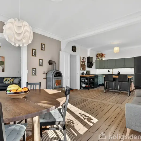 Spisebord med frugtskål står i et lyst køkken-alrum. Et rundt bord er omgivet af stole. I baggrunden ses et køkken med mørke skabe og en grå sofa.