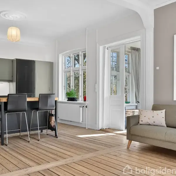 Seng med grøn sengetæppe står i et soveværelse med skråloft. Ved siden af er et natbord med en lampe og plante. Sollys strømmer ind gennem et vindue med gardiner.
