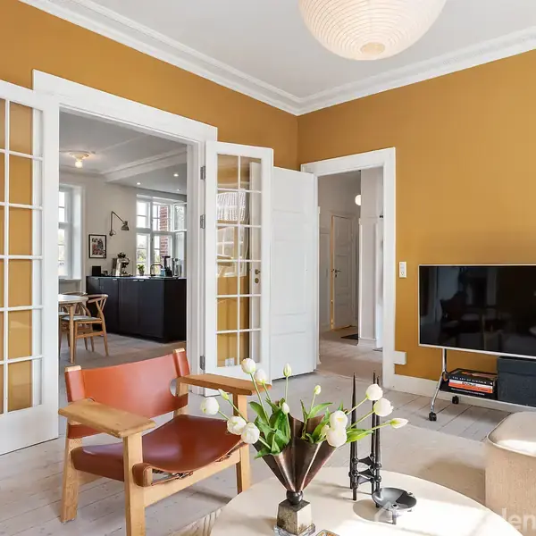 Stue med moderne møblering; en lænestol ved siden af et sofabord med en vase blomster. En fladskærms-tv står på en reol. Rummet har gule vægge og glasdobbeltdøre.