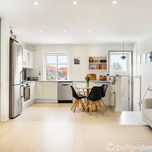 Et moderne køkken i åbent rum forbinder til stue med sofa og fladskærm. Spiseområde i midten har et rundt bord med blomster, omgivet af fire stole.