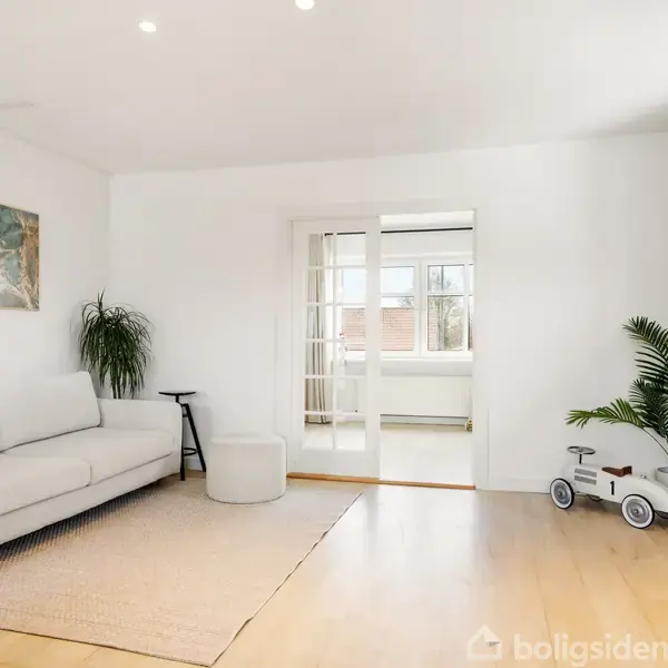 En lys stue med en hvid sofa og sofabord på et tæppe. Ved siden af er der et spisebord med fire sorte stole. Vægdekorationer og en plante skaber en hyggelig atmosfære.