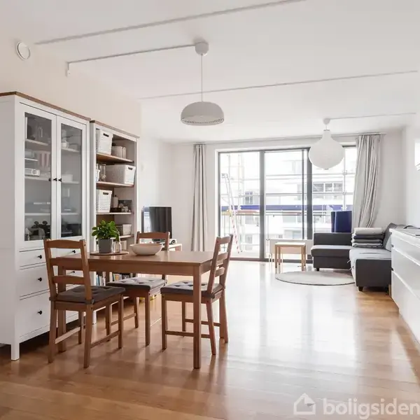 Spisestue med hvidt vitrineskab og træbord med stole. Sofa i baggrunden nær glasdøre, der åbner ud til en altan. Moderne indretning med træ- og hvide elementer.