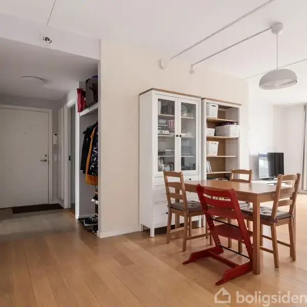 En spisestue med et bord og fire stole, placeret ved siden af et hvidt vitrineskab. Rummet har lyse trægulve og store vinduer til højre, med en åben gang til venstre.