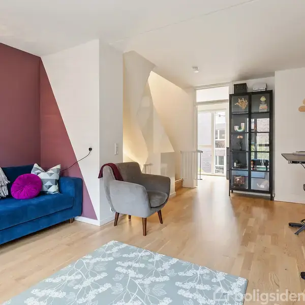 Blå sofa står ved en mørkerød væg med puder; ved siden af står en grå lænestol. Et skrivebord med computer ses til højre. Plankegulv og rummets udsigt mod en balkon.