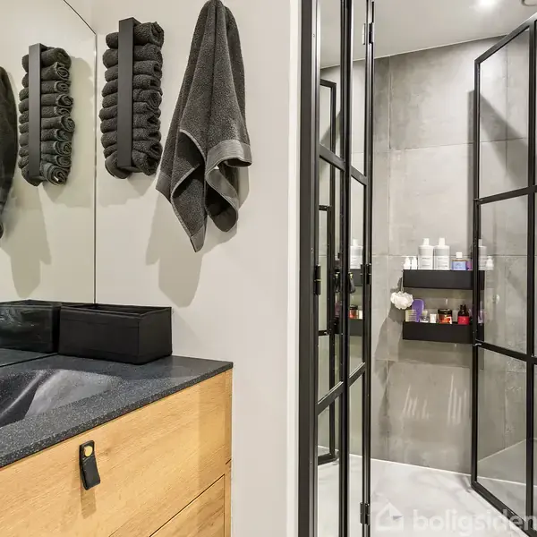 Badeværelse med sort vask og træskabe, håndklæder hængende på væggen, glasdør i metalramme fører til bruseniche med fliser. Toiletries er placeret på hylder i brusenichen.