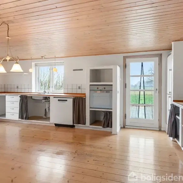 Køkken med træloft og trægulv, indeholder komfur og vask i skab med gardinforhæng, omgivet af vinduer og en glasdør, der fører ud til en grøn have.