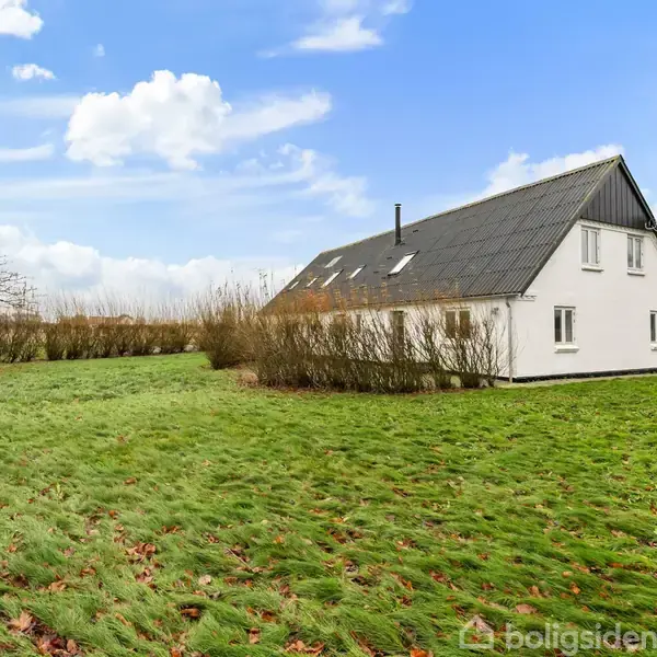 Hvidt hus med sort tag står stille på en stor græsplæne omgivet af nøgne træer og buske under en blå himmel med skyer.